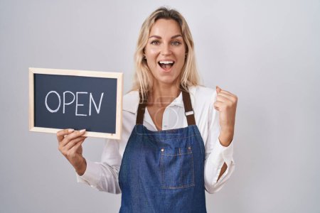 Photo for Young blonde woman wearing apron holding blackboard with open word screaming proud, celebrating victory and success very excited with raised arms - Royalty Free Image