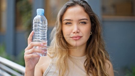 Foto de Joven mujer hispana hermosa sosteniendo botella de agua con expresión relajada en la calle - Imagen libre de derechos