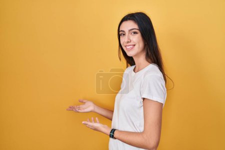 Foto de Joven hermosa mujer de pie sobre fondo amarillo invitando a entrar sonriente natural con la mano abierta - Imagen libre de derechos