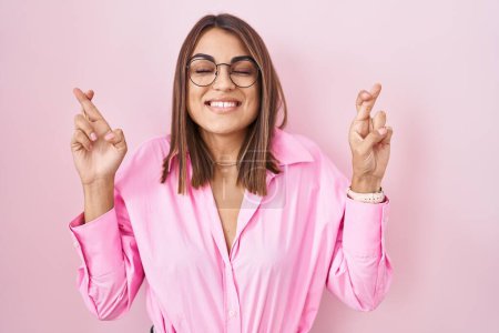 Téléchargez les photos : Jeune femme hispanique portant des lunettes debout sur fond rose geste doigt croisé souriant avec espoir et les yeux fermés. chance et concept superstitieux. - en image libre de droit
