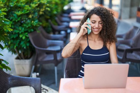 Téléchargez les photos : Jeune belle femme hispanique parlant sur smartphone en utilisant un ordinateur portable assis sur la table à la terrasse du café - en image libre de droit