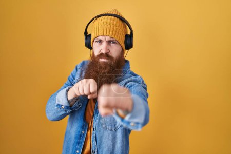 Téléchargez les photos : Homme caucasien à longue barbe écoutant de la musique à l'aide d'écouteurs frappant poing pour se battre, attaque agressive et en colère, menace et violence - en image libre de droit