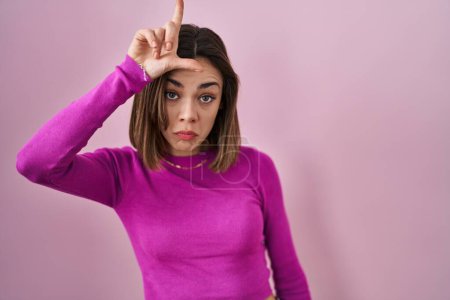 Photo for Hispanic woman standing over pink background making fun of people with fingers on forehead doing loser gesture mocking and insulting. - Royalty Free Image