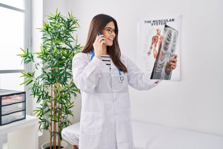 Téléchargez les photos : Jeune belle hispanique femme médecin parler sur smartphone tenant des rayons X à la clinique - en image libre de droit