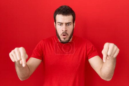 Foto de Joven hombre hispano con camiseta roja casual apuntando hacia abajo con los dedos mostrando publicidad, cara sorprendida y boca abierta - Imagen libre de derechos
