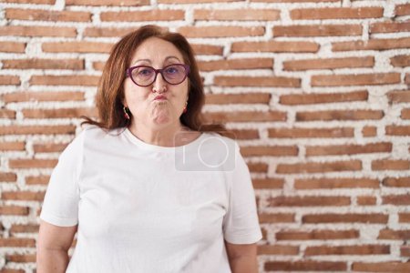 Foto de Mujer mayor con gafas de pie sobre ladrillos pared hinchando mejillas con cara divertida. boca hinchada de aire, expresión loca. - Imagen libre de derechos