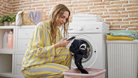 Foto de Mujer rubia joven lavando ropa usando smartphone sonriendo en la sala de lavandería - Imagen libre de derechos