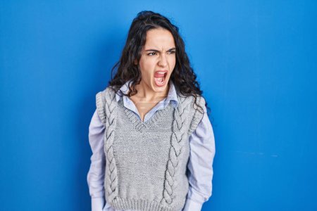 Photo for Young brunette woman standing over blue background angry and mad screaming frustrated and furious, shouting with anger. rage and aggressive concept. - Royalty Free Image