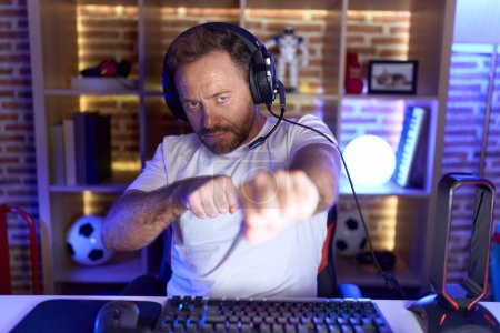 Photo for Middle age man with beard playing video games wearing headphones punching fist to fight, aggressive and angry attack, threat and violence - Royalty Free Image