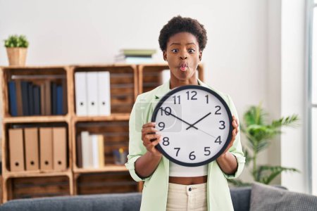 Photo for African american woman holding big clock making fish face with mouth and squinting eyes, crazy and comical. - Royalty Free Image