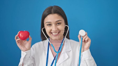 Foto de Joven mujer hispana hermosa doctora sonriendo confiada sosteniendo estetoscopio y corazón sobre fondo azul aislado - Imagen libre de derechos