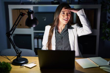 Photo for Young brunette woman working at the office at night with laptop very happy and smiling looking far away with hand over head. searching concept. - Royalty Free Image