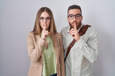 Téléchargez les photos : Jeune couple debout sur fond blanc demandant de se taire avec le doigt sur les lèvres. silence et concept secret. - en image libre de droit