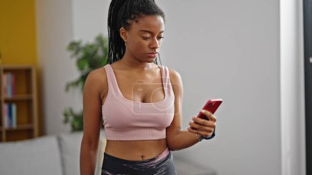 Téléchargez les photos : Femme afro-américaine portant des vêtements de sport en utilisant un smartphone avec une expression sérieuse à la maison - en image libre de droit