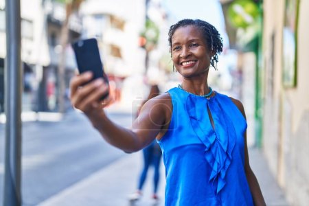Téléchargez les photos : Moyen Age femme afro-américaine souriant confiant faire selfie par le smartphone à la rue - en image libre de droit