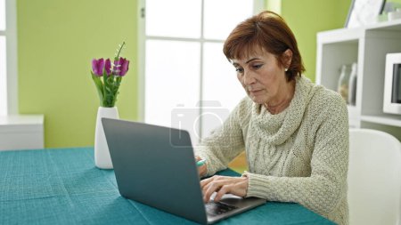 Téléchargez les photos : Femme hispanique mature tenant respirateur vape assis sur la table en utilisant un ordinateur portable à la salle à manger - en image libre de droit
