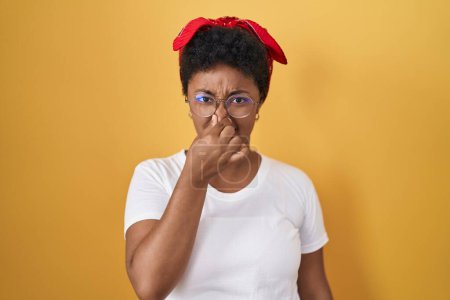 Téléchargez les photos : Jeune femme afro-américaine debout sur fond jaune sentant quelque chose de puant et dégoûtant, odeur intolérable, retenant souffle avec les doigts sur le nez. mauvaise odeur - en image libre de droit