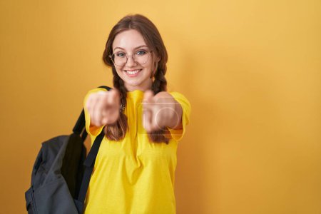 Photo for Young caucasian woman wearing student backpack over yellow background pointing to you and the camera with fingers, smiling positive and cheerful - Royalty Free Image