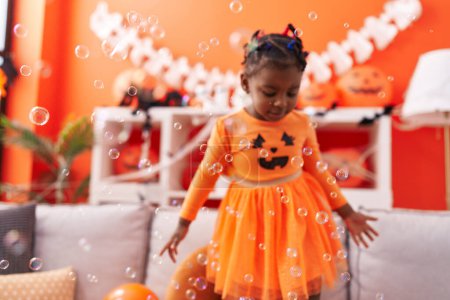 Photo for African american girl wearing pumpkin costume playing with soap bubbles at home - Royalty Free Image