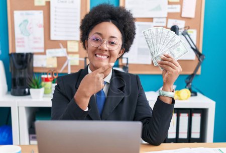 Photo for Beautiful african woman with curly hair holding money at the office smiling happy pointing with hand and finger - Royalty Free Image