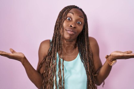 Foto de Mujer afroamericana de pie sobre fondo rosa expresión despistada y confusa con los brazos y las manos levantadas. concepto de duda. - Imagen libre de derechos