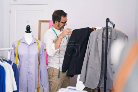Foto de Hombre de mediana edad sastre sonriendo confiado sosteniendo la ropa en el estante en sastrería - Imagen libre de derechos