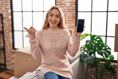 Téléchargez les photos : Femme hispanique se déplaçant vers une nouvelle maison montrant écran smartphone pointant pouce vers le côté souriant heureux avec bouche ouverte - en image libre de droit