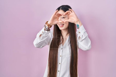Photo for Chinese young woman standing over pink background doing heart shape with hand and fingers smiling looking through sign - Royalty Free Image