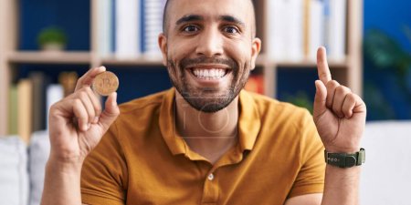 Téléchargez les photos : Homme hispanique avec barbe tenant monnaie virtuelle Bitcoin sourire avec une idée ou une question pointant du doigt avec un visage heureux, numéro un - en image libre de droit