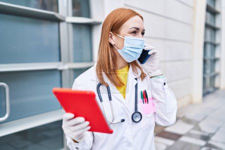 Foto de Mujer joven caucásica médico con máscara médica usando touchpad hablando en el teléfono inteligente en el hospital - Imagen libre de derechos