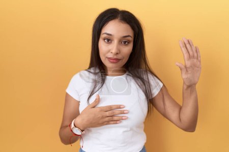 Foto de Mujer árabe joven con camiseta blanca casual sobre fondo amarillo jurando con la mano en el pecho y la palma abierta, haciendo un juramento de lealtad promesa - Imagen libre de derechos