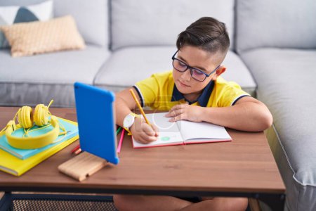 Photo for Adorable hispanic boy drawing on book looking touchpad at home - Royalty Free Image