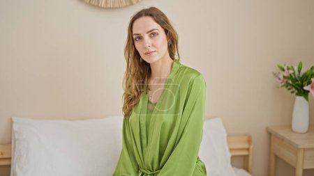 Téléchargez les photos : Jeune femme blonde assise sur le lit avec un visage sérieux dans la chambre - en image libre de droit