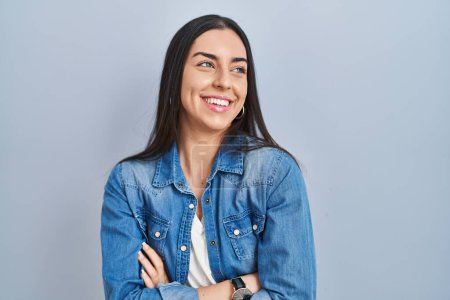 Foto de Mujer hispana de pie sobre fondo azul mirando hacia otro lado con sonrisa en la cara, expresión natural. riendo confiado. - Imagen libre de derechos