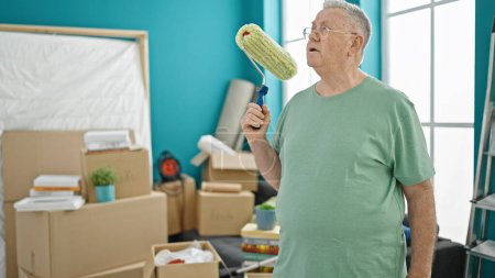Foto de Hombre de pelo gris de mediana edad sonriendo confiado sosteniendo el rodillo de pintura en un nuevo hogar - Imagen libre de derechos