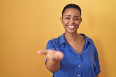 Photo for African american woman standing over yellow background smiling cheerful offering palm hand giving assistance and acceptance. - Royalty Free Image