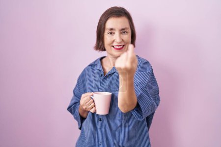 Foto de Mujer hispana de mediana edad bebiendo una taza de café haciendo señas ven aquí gesto con la mano invitando a la bienvenida feliz y sonriente - Imagen libre de derechos