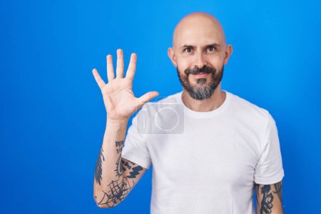 Téléchargez les photos : Homme hispanique avec des tatouages debout sur fond bleu montrant et pointant vers le haut avec les doigts numéro cinq tout en souriant confiant et heureux. - en image libre de droit