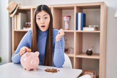 Photo for Chinese young woman putting coin in piggy bank scared and amazed with open mouth for surprise, disbelief face - Royalty Free Image