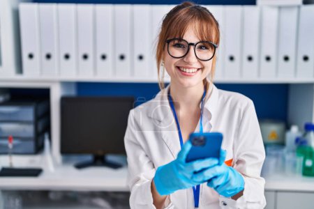 Téléchargez les photos : Jeune femme scientifique souriant confiant en utilisant un smartphone au laboratoire - en image libre de droit