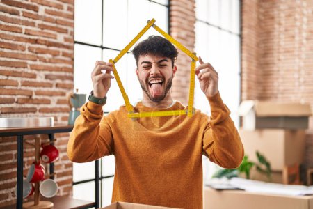 Foto de Hombre hispano con barba mudándose a un nuevo hogar sosteniendo la regla sacando la lengua feliz con expresión divertida. - Imagen libre de derechos