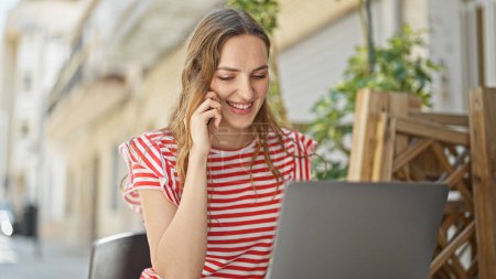 Téléchargez les photos : Jeune femme blonde parlant sur smartphone en utilisant un ordinateur portable assis sur la table à la terrasse du café - en image libre de droit