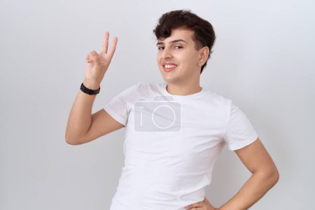 Téléchargez les photos : Jeune homme non binaire portant un t-shirt blanc décontracté souriant regardant la caméra montrant des doigts faisant signe de victoire. numéro deux. - en image libre de droit