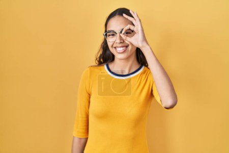 Téléchargez les photos : Jeune femme brésilienne portant des lunettes sur fond jaune souriant heureux faire ok signe avec la main sur les yeux regardant à travers les doigts - en image libre de droit