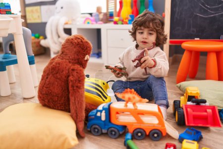 Téléchargez les photos : Adorable garçon hispanique jouant avec voiture et jouet de dinosaure assis sur le sol à la maternelle - en image libre de droit