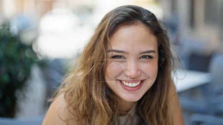 Foto de Joven hermosa mujer hispana sonriendo confiada sentada en la mesa en la terraza de la cafetería - Imagen libre de derechos