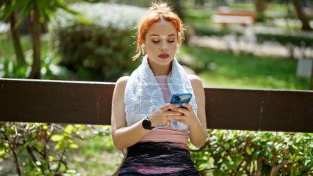 Téléchargez les photos : Jeune rousse femme en utilisant smartphone et écouteurs assis sur le banc au parc - en image libre de droit
