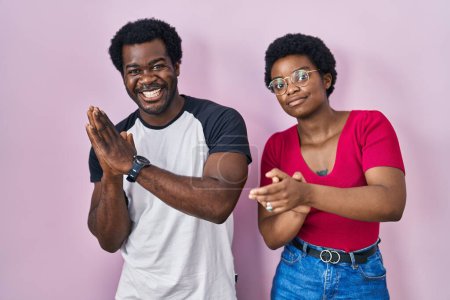 Téléchargez les photos : Jeune couple afro-américain debout sur fond rose applaudissant heureux et joyeux, souriant mains fières ensemble - en image libre de droit