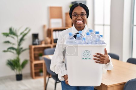 Foto de Mujer afroamericana trabajando en la oficina sosteniendo una botella de plástico para reciclar sonriendo y riendo en voz alta porque divertida broma loca. - Imagen libre de derechos