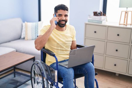 Foto de Hombre árabe joven hablando en el teléfono inteligente sentado en silla de ruedas en casa - Imagen libre de derechos